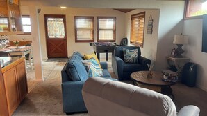 Un salon chaleureux avec un canapé bleu, un fauteuil blanc et une table basse. On y trouve également une table à manger en bois avec des chaises, un baby-foot et une porte qui donne sur une autre pièce.