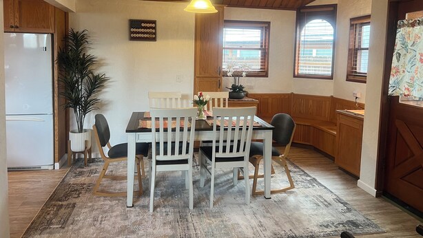 Un coin repas avec une table en bois, des chaises blanches et un lustre. Un réfrigérateur se trouve à gauche, et une fenêtre avec des stores est visible au fond.
