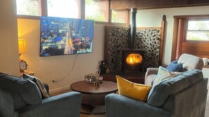 Un salon chaleureux avec deux fauteuils bleus, une table basse ronde en bois, une cheminée en pierre, un téléviseur fixé au mur et une fenêtre donnant sur des immeubles.