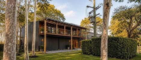 Une maison moderne à deux étages, dotée d’un balcon, entourée d’arbres et d’une pelouse bien entretenue.