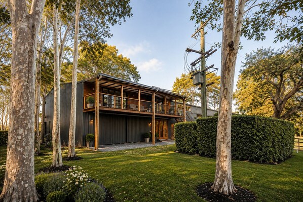 A modern two-story house with a balcony, surrounded by trees and a well-maintained lawn.
