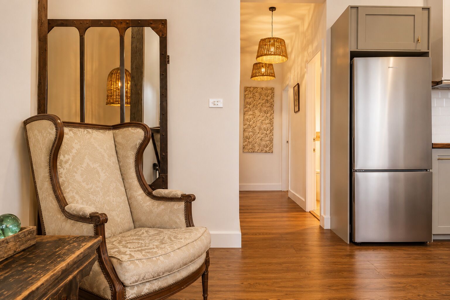 A room with a wooden chair, a wooden table, a modern stainless steel refrigerator, and a hallway with a patterned wall and hanging lights.