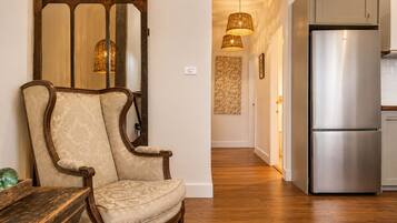A room with a wooden chair, a wooden table, a modern stainless steel refrigerator, and a hallway with a patterned wall and hanging lights.