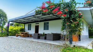 A covered outdoor seating area with a table and chairs, a gravel driveway, and a house with a white exterior and green shutters. - Chalet - private family estate near Parque del Café - Coffee Region. (Quimbaya)