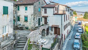 A narrow street with traditional stone buildings, a small outdoor seating area with tables and chairs, and parked cars along the side. - Apartment Viola by Interhome (Gello)