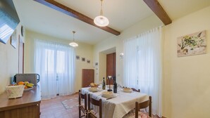A dining area with a table set for four, wooden chairs, a kitchen counter with fruit, and a painting on the wall. - Apartment Viola by Interhome (Gello)
