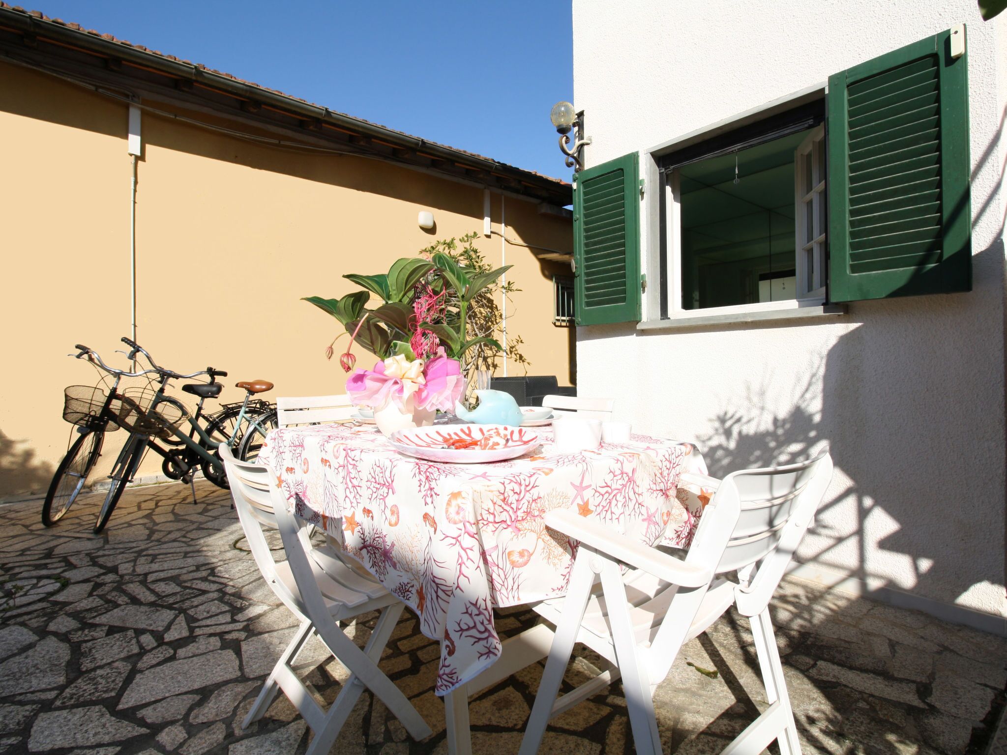 Un patio con una mesa dispuesta para dos, rodeado de sillas blancas, una bicicleta y una maceta.