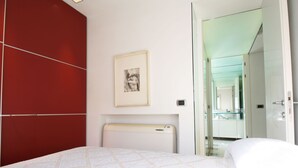 Un dormitorio moderno con cama, pared de acento roja, un cuadro enmarcado y un baño visible a través de una puerta abierta.