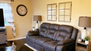 Una sala de estar con un sofá de cuero marrón oscuro, una mesa de centro negra, dos lámparas de pared, un reloj y una ventana con cortinas.