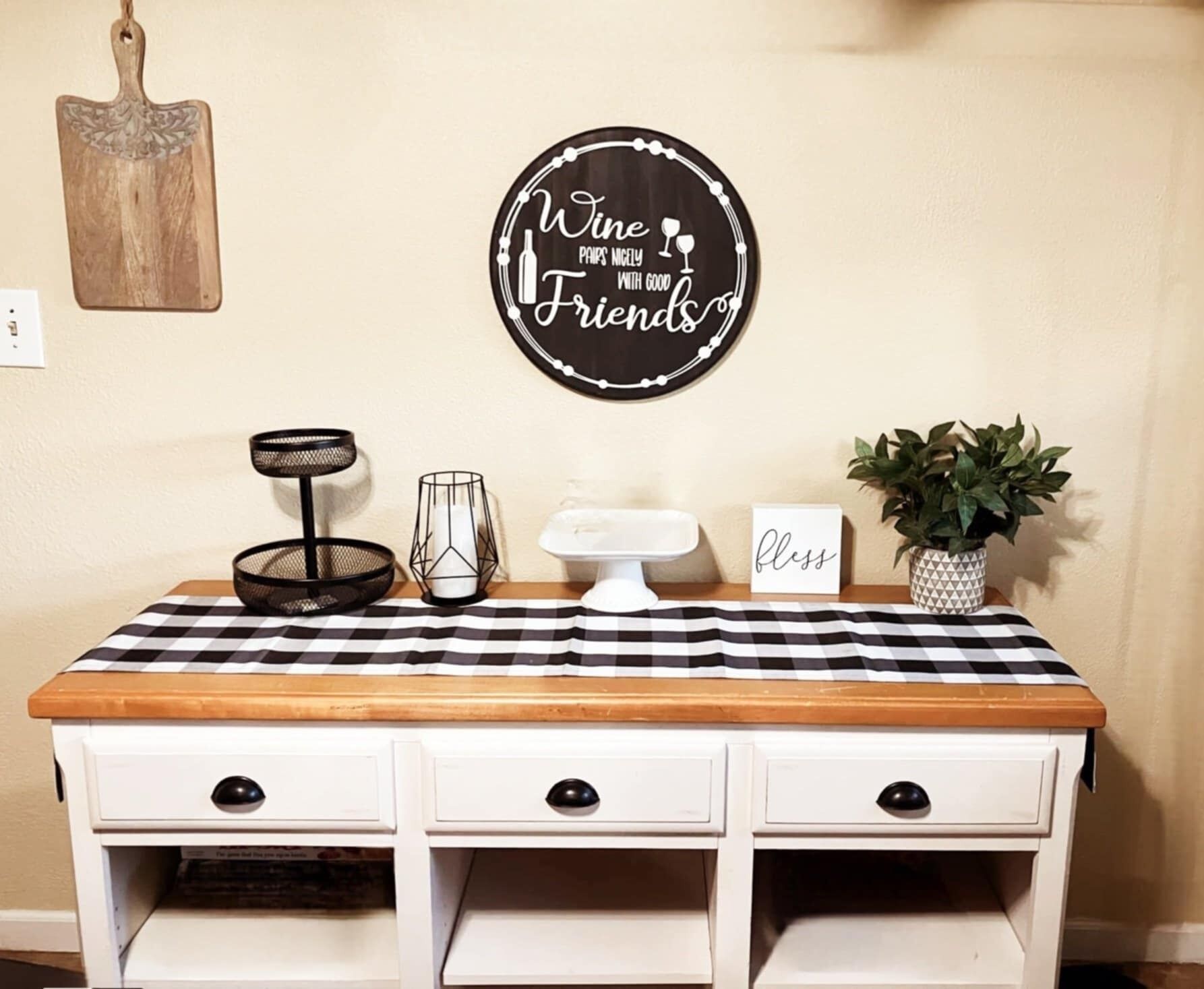 Uma bancada de cozinha com toalha de mesa xadrez, um vaso de planta, um castiçal e uma placa decorativa na parede.