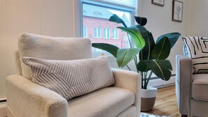 A plush armchair with a textured cushion, positioned next to a large potted plant with broad leaves, in a room with a view of a red building through a window. - Newly Refreshed 3bd Spacious Unit Minutes from 290 (Worcester)