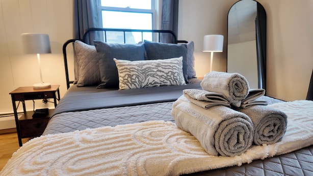 A neatly made bed with rolled towels, a bedside table with a lamp, and a large mirror on the wall. - Newly Refreshed 3bd Spacious Unit Minutes from 290 (Worcester)