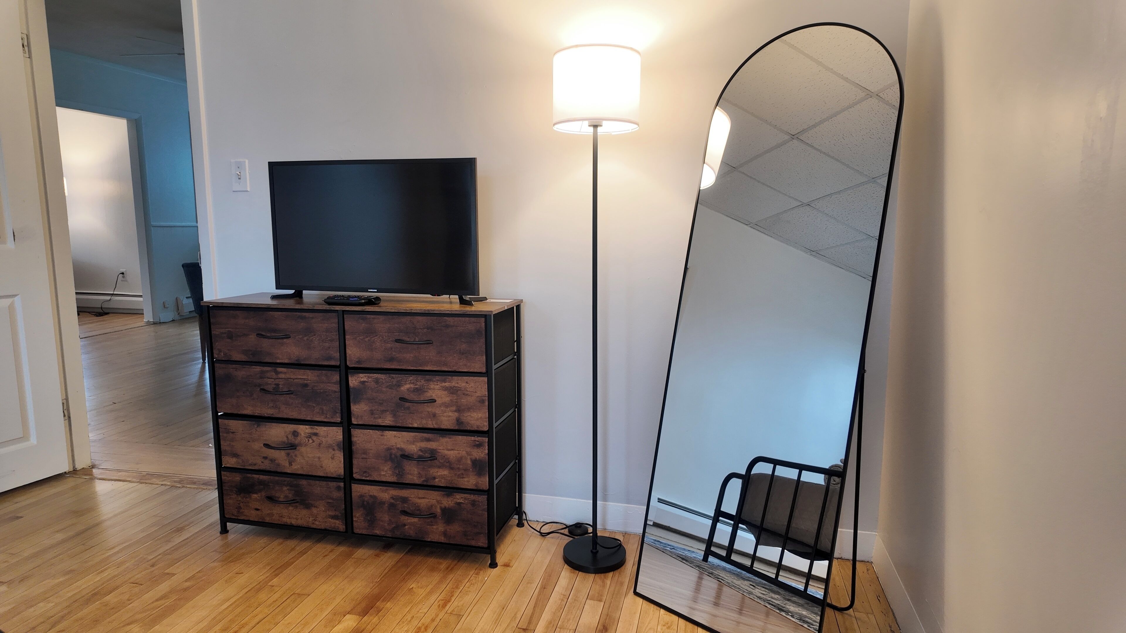 Een kamer met een houten dressoir, een flatscreen televisie, een vloerlamp en een grote spiegel.
