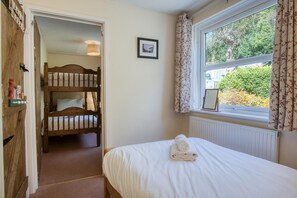 A bedroom with a single bed, a window with curtains, a radiator, and a wooden door leading to a bunk bed area.