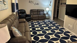 A living room with a ceiling fan, two leather sofas, a patterned rug, and a wall with a 'Relax at the Lake' sign.