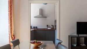 A modern kitchen with a stainless steel range hood, a dining table with a fruit bowl, and a TV on a cabinet. - GuestHost - Rione Prati Apartment A (Rome)