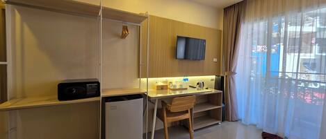 A hotel room with a kitchenette, a TV mounted on the wall, a desk with a chair, and a large window with curtains.