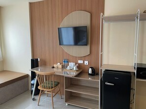 Een compacte hotelkamer met een wand van houten panelen, een flatscreen televisie boven een houten bureau, een kleine koelkast en een stoel met kussen.