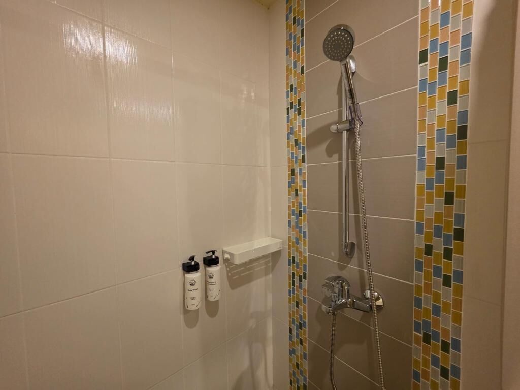 A shower with a handheld showerhead and a wall-mounted faucet, a tiled wall with a colorful border, and a white shelf with toiletries.