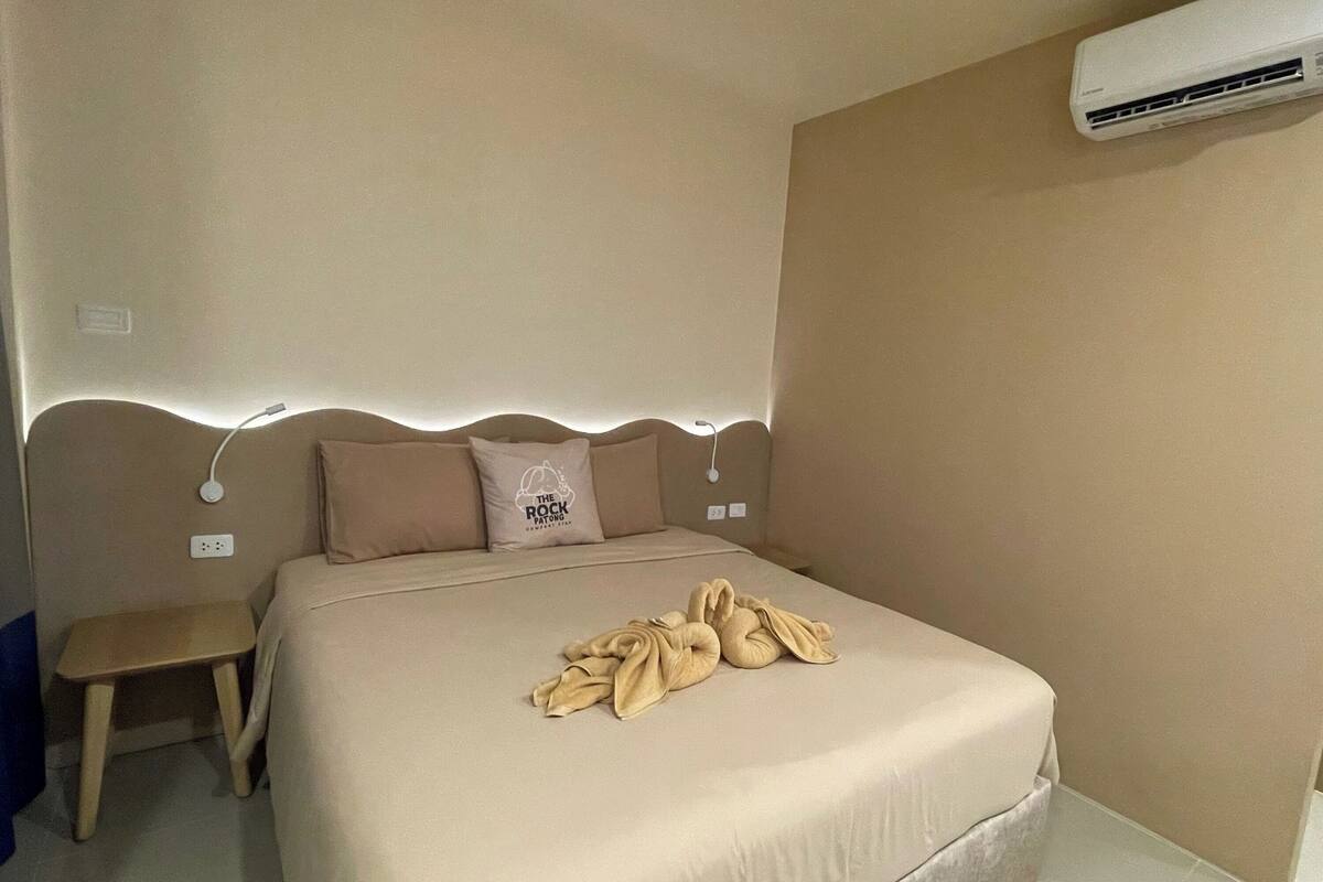 Una cama bien hecha con una manta y almohadas color beige, una mesita de noche con lámpara y un equipo de aire acondicionado en la pared.