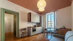A modern living room with a sofa, dining table, and chairs. There is a kitchen area with cabinets and a stove. The room has wooden flooring and a large window with curtains. - GuestHost - Rione Prati Apartment B (Rome)