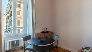 A corner room with a round table, two chairs, a radiator, a small cabinet, and a window with a view of buildings. - GuestHost - Rione Prati Apartment B (Rome)