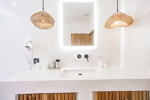 A modern bathroom with a white sink, two wicker pendant lights, a mirror, and wooden cabinets below the sink. - Villa Calipso (Gili Trawangan)