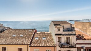 A coastal cityscape with terracotta-roofed buildings, a clear sky, and the sea in the background.