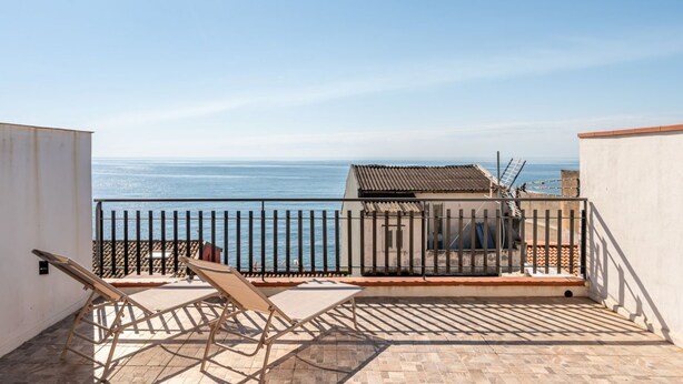 A balcony with a view of the sea, two lounge chairs, and a small building with a tiled roof.
