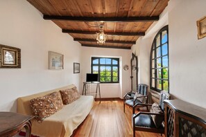 A room with a wooden ceiling, a bed, a sofa, a desk, a chair, and a window with a view of greenery.
