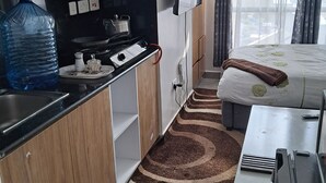 A compact hotel room with a kitchenette, a large water dispenser, a flat-screen TV, a bed with a floral duvet, and a view of the city through large windows. - RnM Haven in Tsavo Apartments 13kms from JKIA
Walk to Stylish,Clean & Comfy Stay (Nairobi)