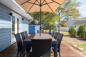 Eine Terrassengruppe mit Tisch und Stühlen, ein großer Sonnenschirm und ein blauer Schuppen im Hintergrund.