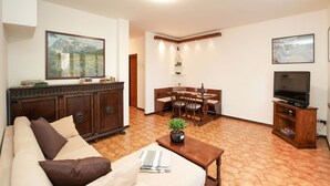 Een woonkamer met een bank, een salontafel met een plant, een eethoek met een tafel en stoelen, een televisie op een houten standaard, en een schilderij aan de muur.