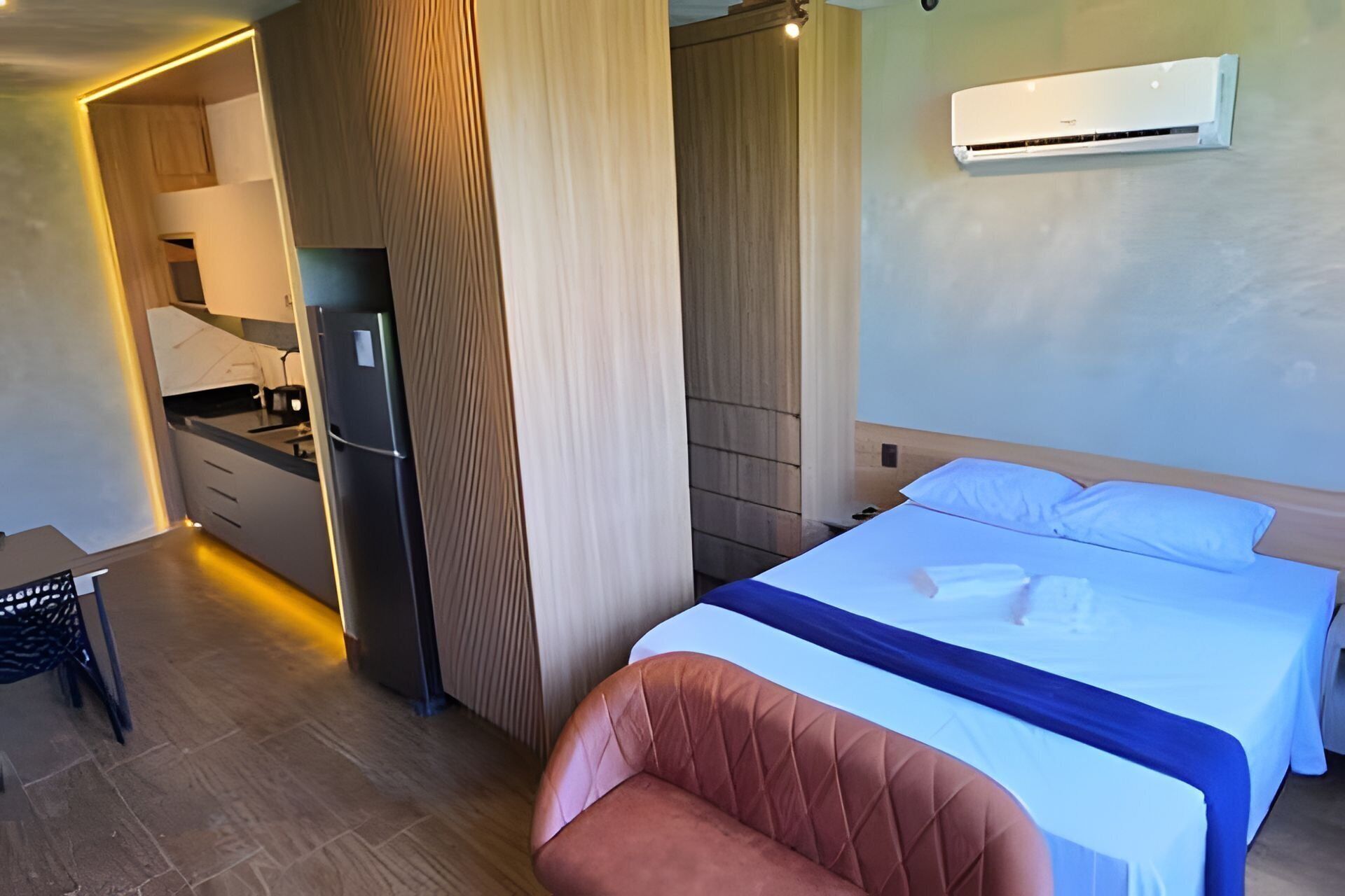 A hotel room with a bed, a chair, a kitchenette, and an air conditioning unit.