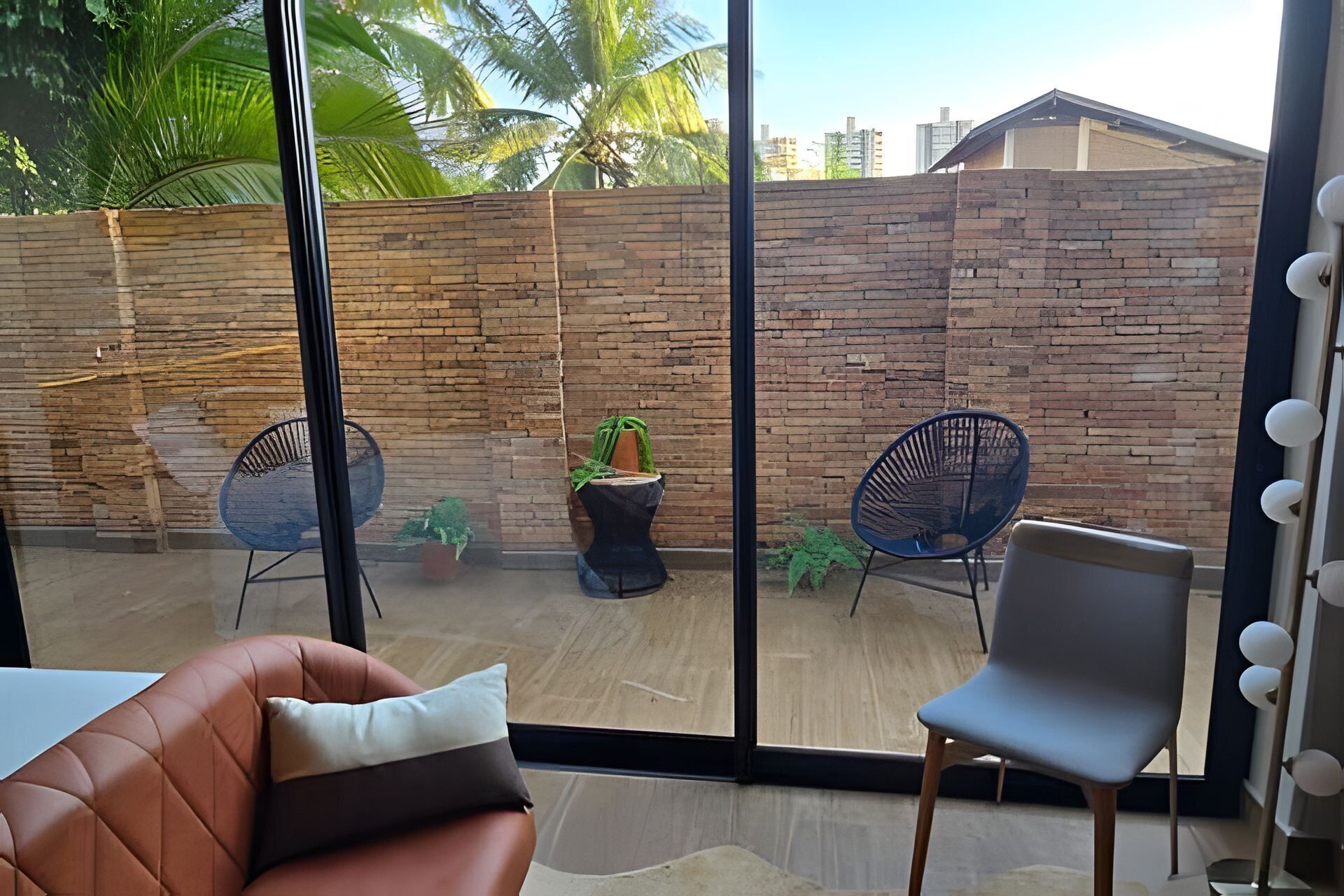 A modern room with a large sliding glass door leading to an outdoor patio. The patio features a brick wall, two black metal chairs, and a small table with a potted plant.