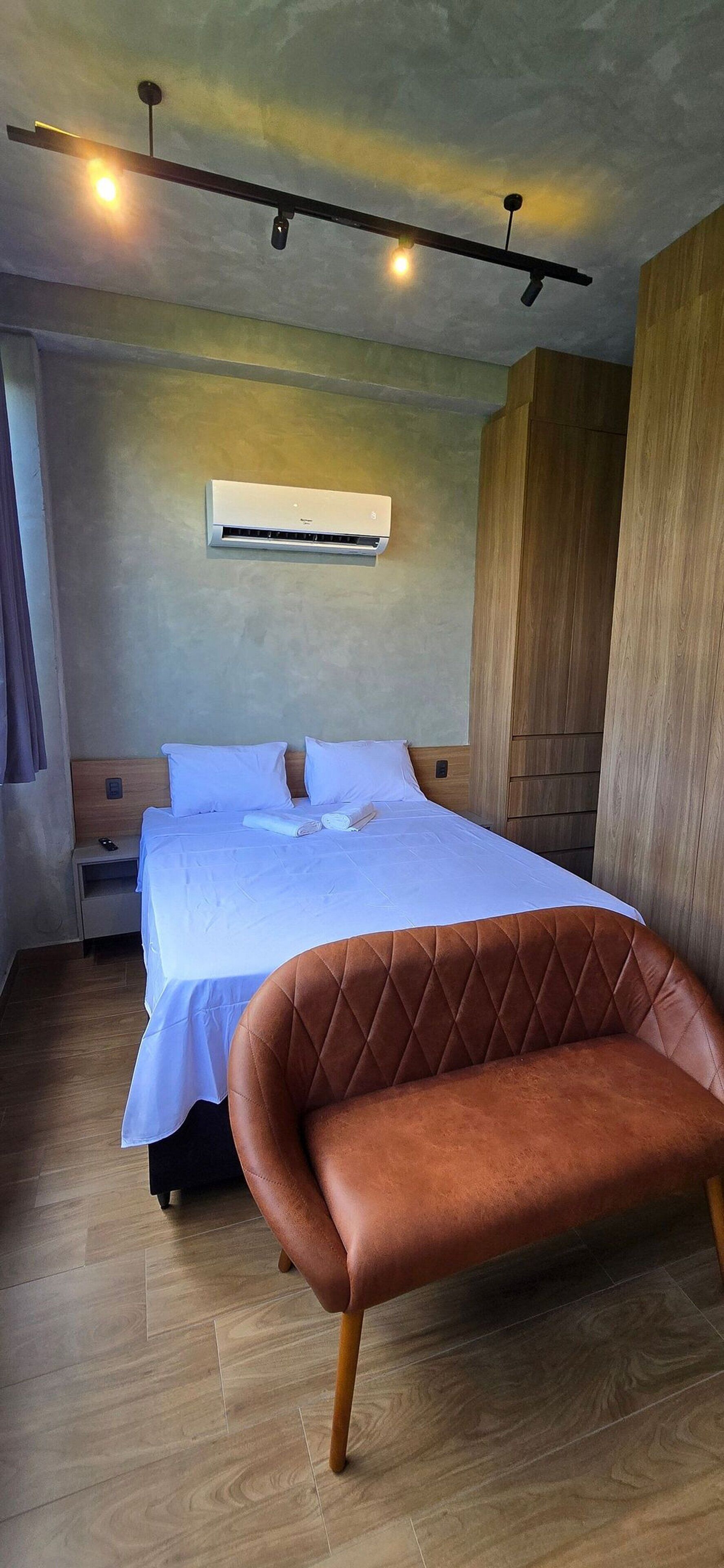 Ein Hotelzimmer mit einem Bett, einem braunen Ledersessel, einem Holzschrank, einem Nachttisch und einer Klimaanlage.