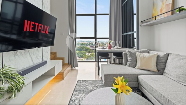 A modern living room with a large flat-screen TV displaying the Netflix logo, a grey sofa, a dining area with a table and chairs, and a view of greenery through large windows. - Senso by Kozystay - Tanjung Barat (Jakarta)
