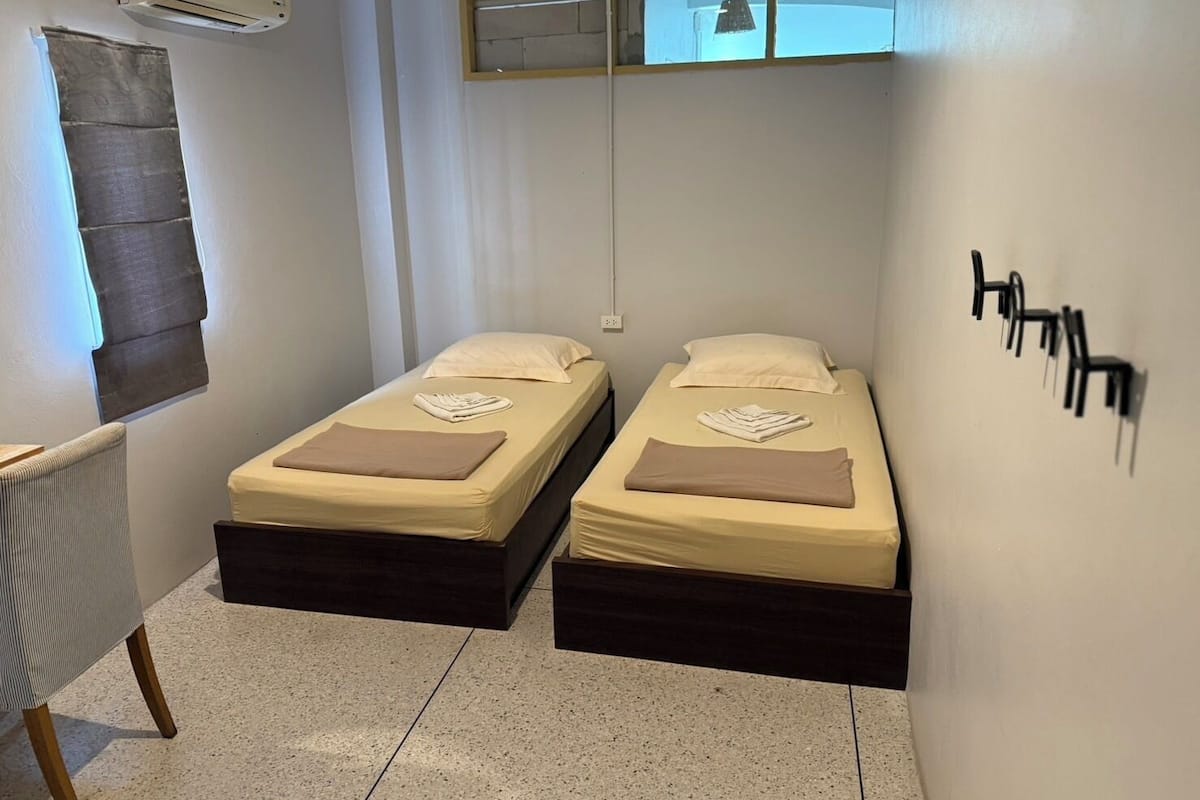 A small, clean room with two single beds, a chair, and a window with blinds.
