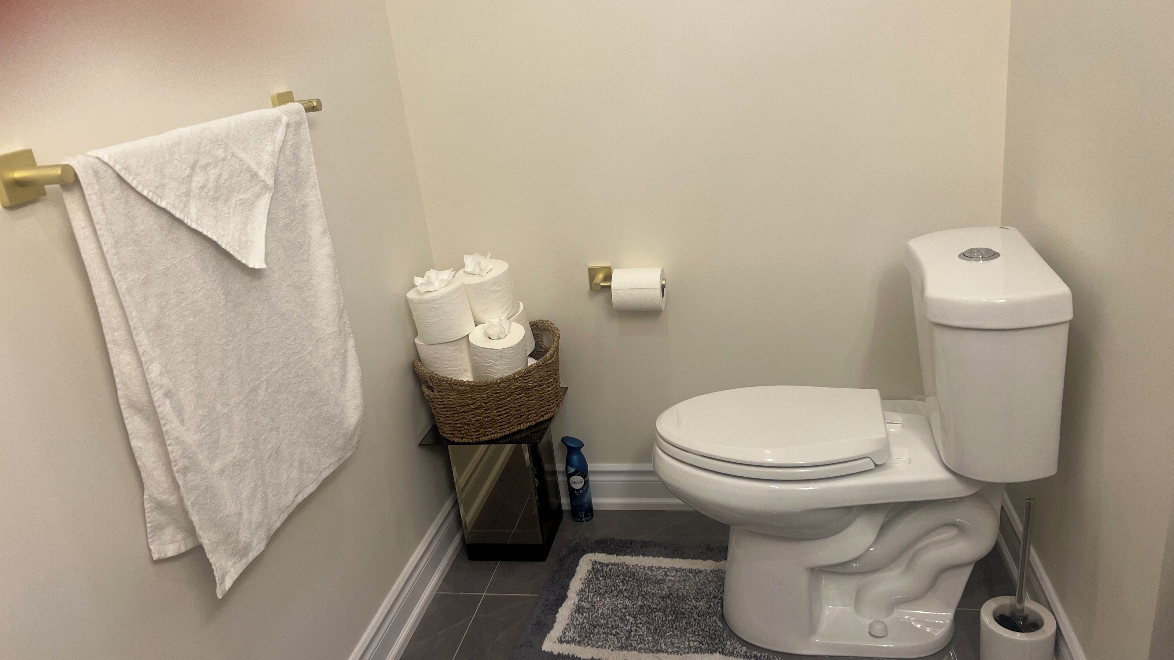 A small bathroom with a toilet, a towel rack with a towel, a basket of toilet paper, and a roll of toilet paper on the wall.