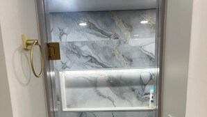 A bathroom with a glass-enclosed shower area, marble walls, and a white shelf with toiletries. - The Luxe Haven (East Gwillimbury)