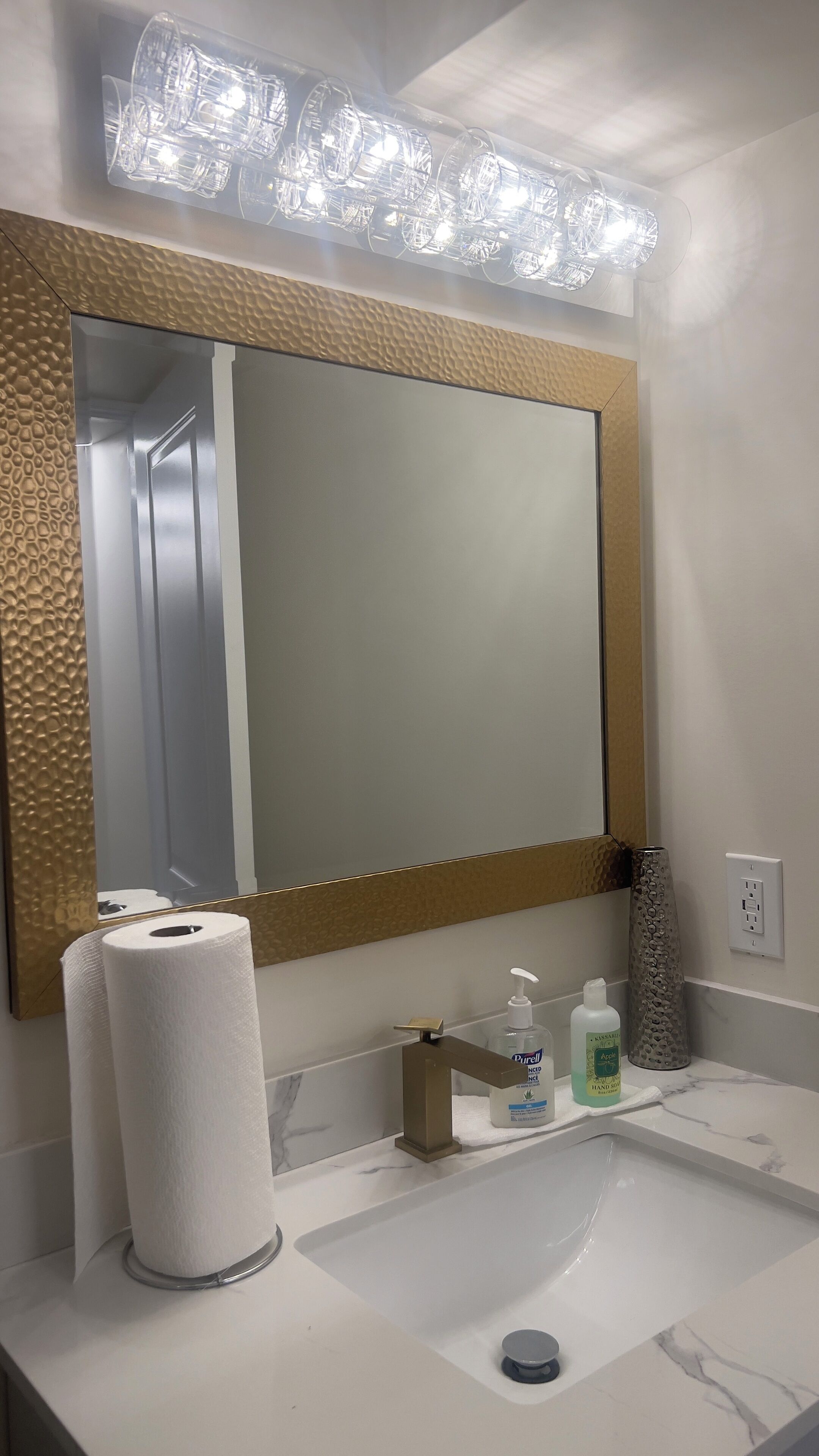 A bathroom with a large mirror, a toilet paper holder, a hand soap dispenser, and a sink.
