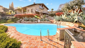 Une piscine dotée d’une terrasse en pierre, entourée de jardins aménagés et d’une maison en pierre.