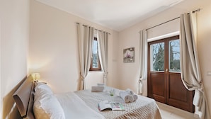 Un lit bien fait, avec un magazine, des serviettes et une petite plante. Il y a une fenêtre avec des rideaux, un tableau au mur et une porte également dotée d’un rideau.
