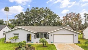 A single-story house with a garage, a front lawn with a small garden, and a large tree with Spanish moss. - Pool Party in Daytona! Sleeps 8 (Holly Hill)