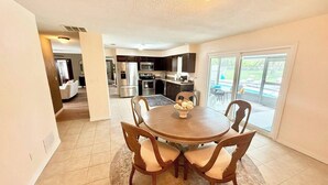 A dining area with a round table and four chairs, a kitchen in the background, and a sliding glass door leading to a balcony. - Pool Party in Daytona! Sleeps 8 (Holly Hill)
