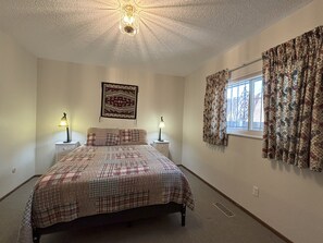 A bedroom with a bed, two bedside tables with lamps, a patterned wall hanging, a window with curtains, and a ceiling light fixture.