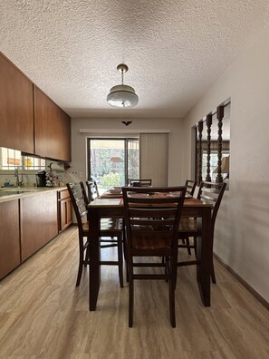 A dining area with a wooden table and chairs, a kitchen with wooden cabinets, and a large window with a view of greenery outside.