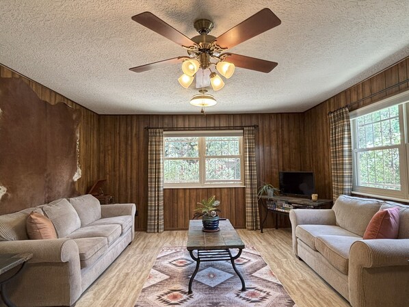 A room with two sofas, a coffee table, a ceiling fan, a TV, and a window with curtains.