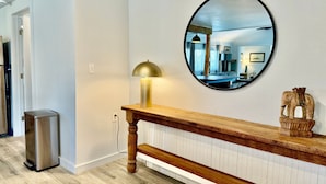 Een hal met een houten bankje, een ronde spiegel, een kleine prullenbak en een lamp.