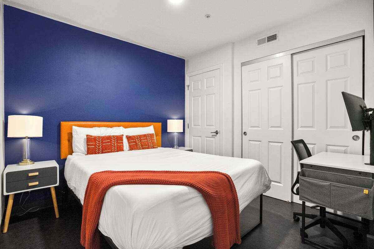 Une chambre d’hôtel avec un lit, un bureau et une chaise. Il y a deux portes et une fenêtre.